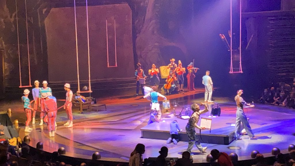 Cirque du Soleil Disney Springs - Preshow the stage is filled with multiple perfomers as they entertain the audience before the show begins. A select number of children that have been invited up from the audience also on stage, each making a drawing on paper as they sit on stage