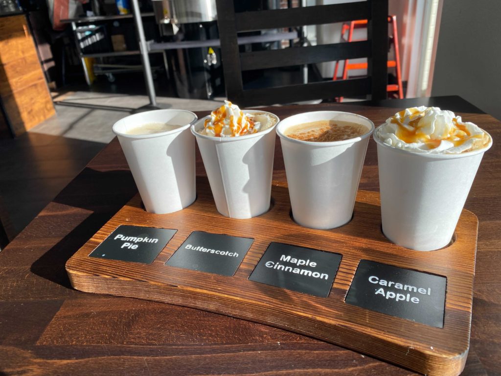 New City Coffee - Coffee Flights offer seasonal flavors year-round. Four paper cups of coffee are places on a wood board that identifies each flavor of coffee. Left to right they are Pumpkin Pie, Butterscotch, Maple Cinnamon, and Caramel Apple