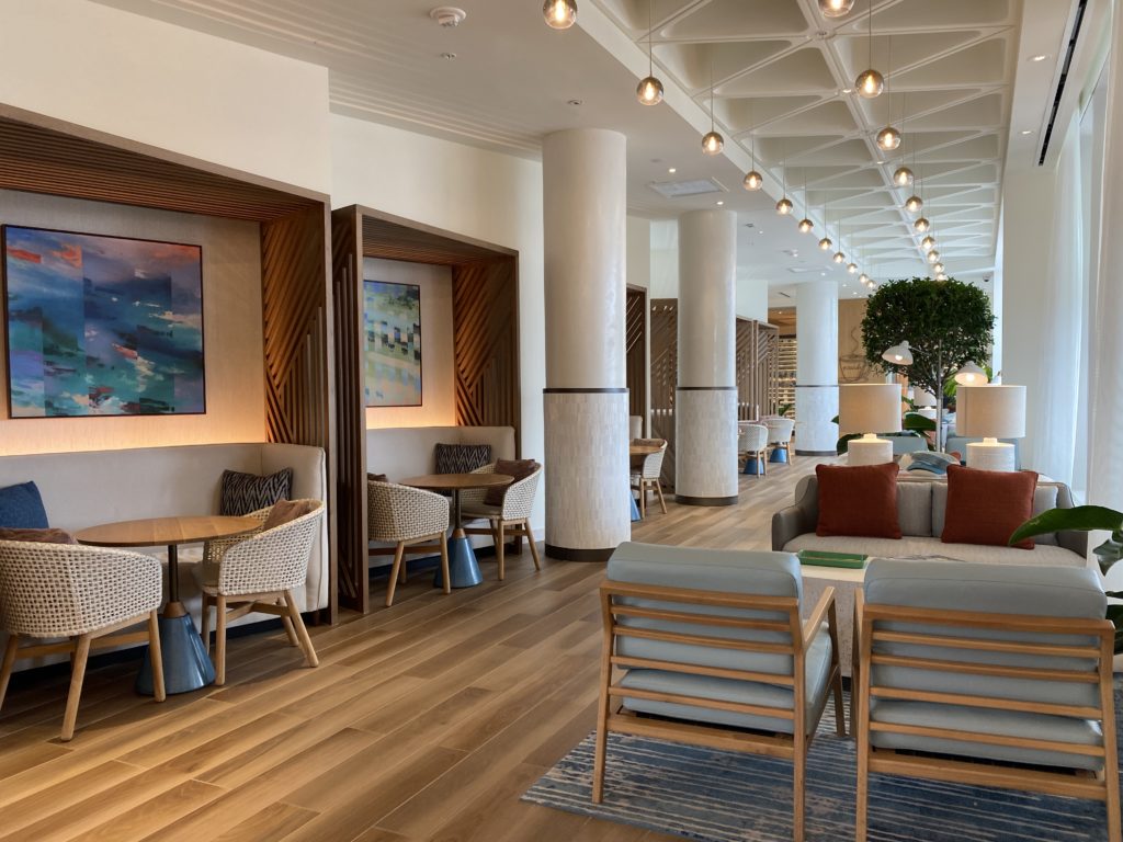 Sitting Area Near Amare at Swan Reserve - multiple tables and chairs with soft beige colored chairs and light brown wood tables line either side of an aisle. The floor is a mix brown wood floor with a bright white ceiling above, with a triangular pattern adding a decorative element to the ceiling. 