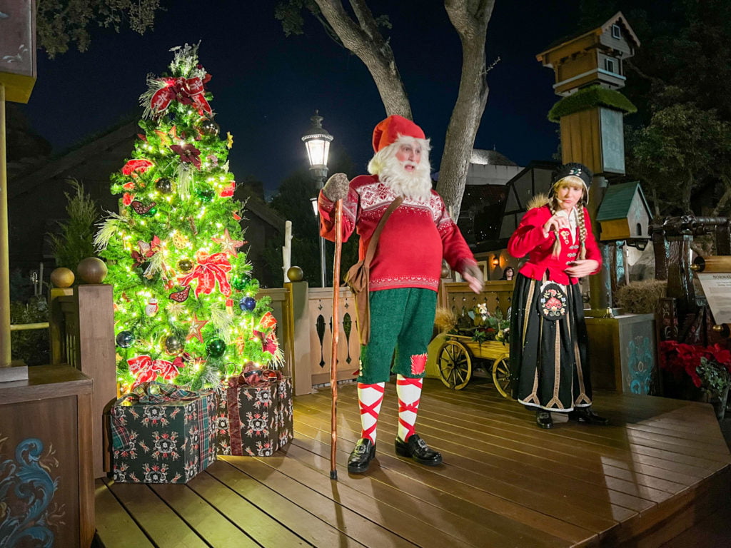 Norway Holiday Storyteller at EPCOT is presented by two storytellers, a male dressed in an elfish looking costume of green shorts, red and white striped socks, and a red sweater, with a white beard and wooden staff. The female storyteller has blonde hair with braids, a red and white sweater, and a long green skirt. They stand on a wooden stage with a Christmas tree on the back with white lights and red ornaments