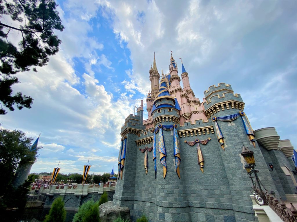 This photo is taken at the wishing well near Cinderella Castle, look up towards the castle which has a gray stone base and pink toned towers and blue turrets on top. There are gold, blue, and purple embellishments on the castle for the 50th anniversary of Walt Disney World. The sky is blue with white puffy clouds.
