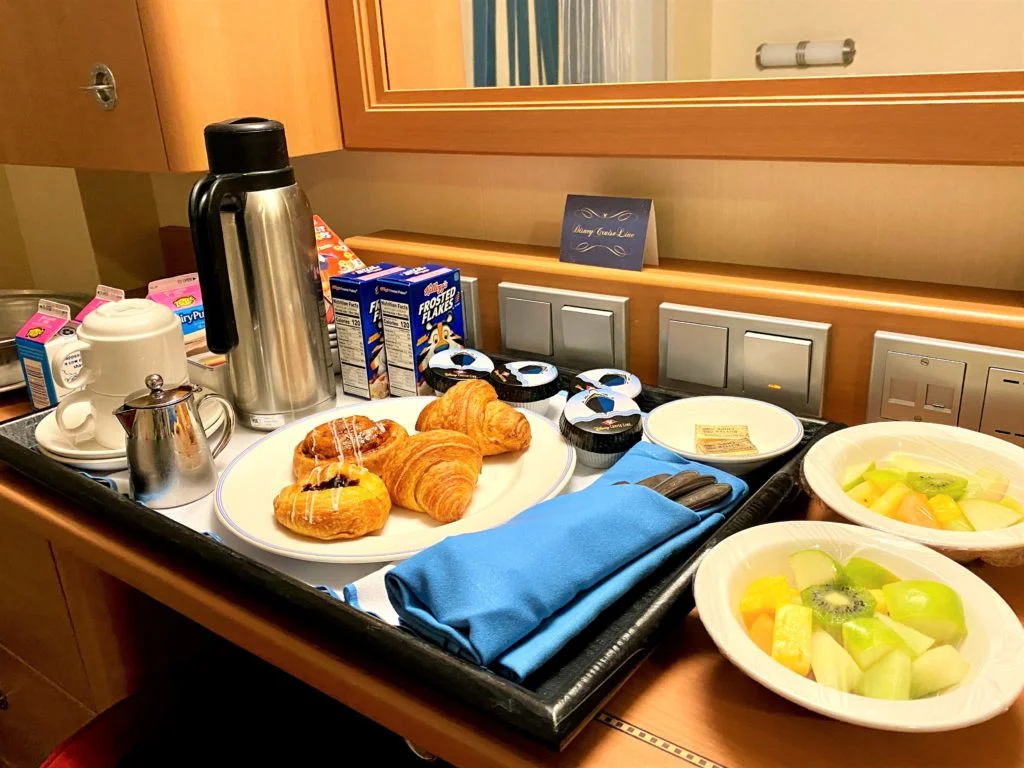 Room Service is included in the price of your Disney Cruise - a plate of Danish pastries and croissants with coffee and fresh fruit sit on a deck in a Disney cruise stateroom
