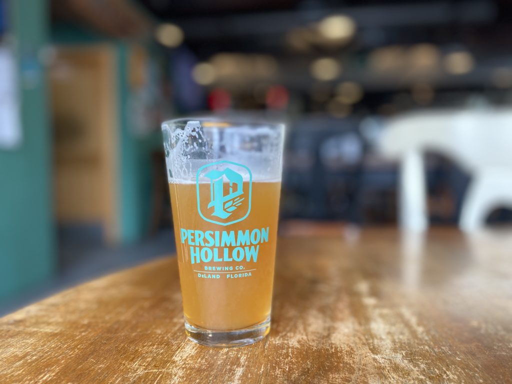 A thick beer glass with the logo of Persimmon Hollow Deland sits on a table