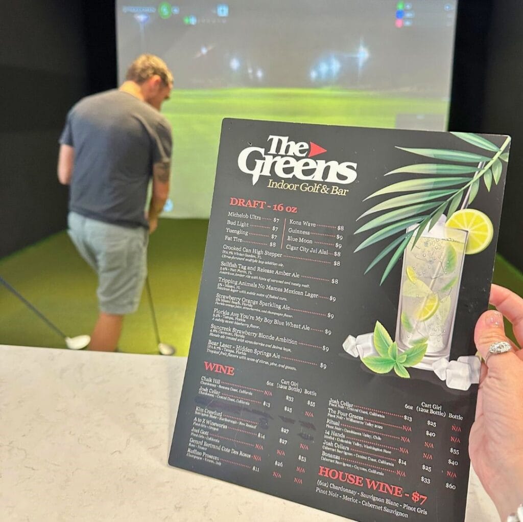 The Greens Indoor Golf and Bar at Pointe Orlando menu in front of camera and man in golf bay - image by Dani Meyering