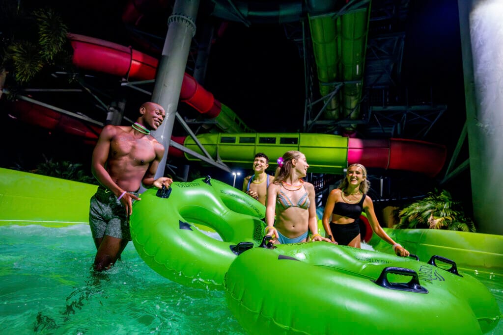 Four young adults in swimsuits stand in a shallow pool at an indoor water park, smiling and chatting while holding onto a large bright green inflatable raft. Colorful enclosed water slides in red and green twist overhead, and the scene is lit with vibrant, neon-like lighting.