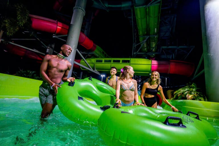 Four young adults in swimsuits stand in a shallow pool at an indoor water park, smiling and chatting while holding onto a large bright green inflatable raft. Colorful enclosed water slides in red and green twist overhead, and the scene is lit with vibrant, neon-like lighting.