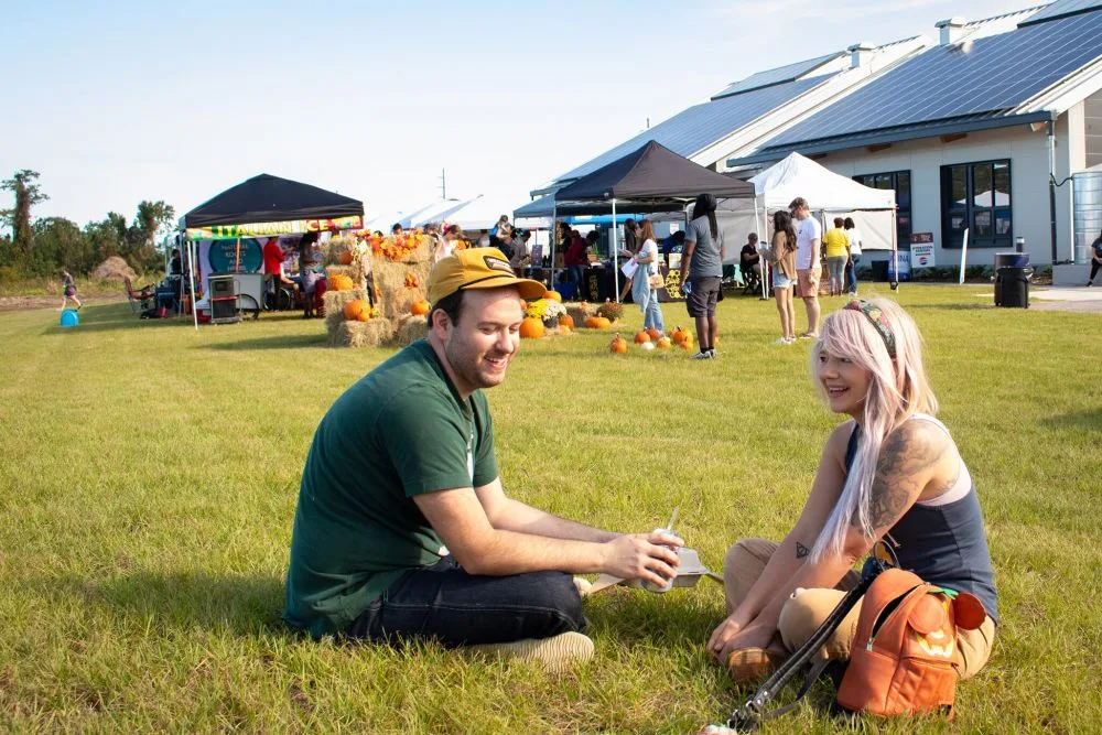A couple on a date sit on the grass smiling with a farm festival going on behind them